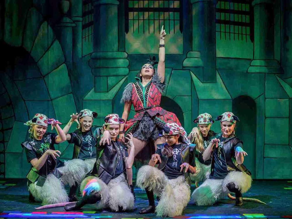 A dynamic stage scene featuring a performer in a striking red and black costume with textured fur details, standing at the centre with one arm confidently pointing upward. Surrounding the performer are a group of children dressed as playful rats, wearing leather vests, furry leg warmers, and bandanas with mouse ears. The vibrant, theatrical backdrop is a stylized dungeon-like setting with arches and columns, illuminated by colourful stage lighting.