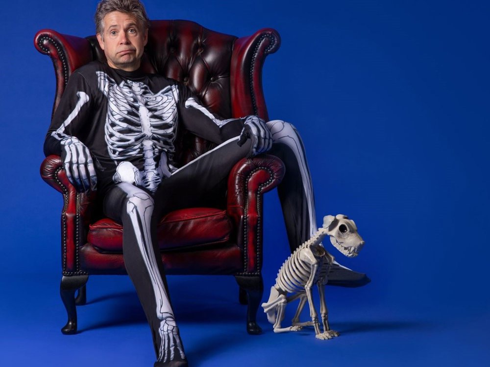 A man wearing a full-body skeleton costume sits in a deep red leather armchair against a solid blue background. He has a slightly surprised expression and tousled hair. Beside him on the floor is a small dog skeleton figure, positioned as if sitting obediently.