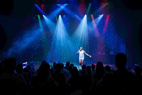 Rikki Beadle-Blair on stage under bright, colourful spotlights, wearing a white sleeveless outfit and holding a microphone. The audience is visible in the foreground, and a DJ stands behind a booth on the right, with a backdrop of twinkling star-like lights.