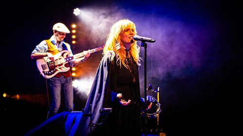 Close-up of a female vocalist with long blonde hair singing into a microphone under purple stage lighting, holding a tambourine. A bass guitarist plays in the background.