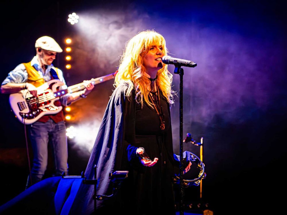 Close-up of a female vocalist with long blonde hair singing into a microphone under purple stage lighting, holding a tambourine. A bass guitarist plays in the background.