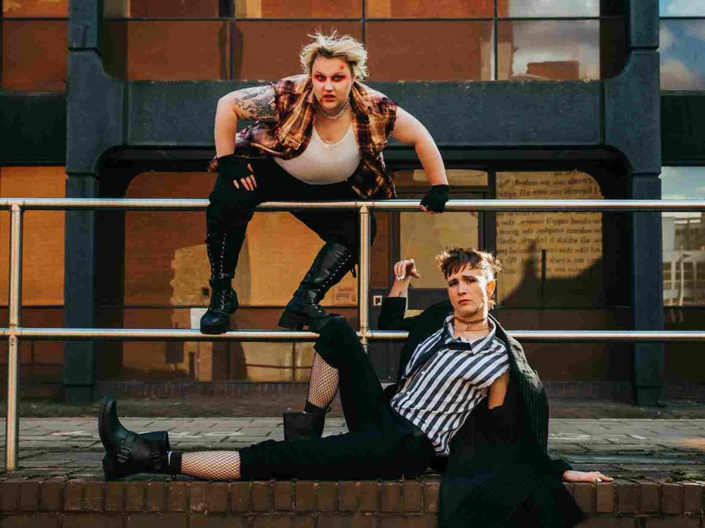 Two people pose outside a modern brick and glass building. One crouches on a railing in a checked shirt and boots, while the other lounges below in a striped shirt, fishnet tights, and long coat.