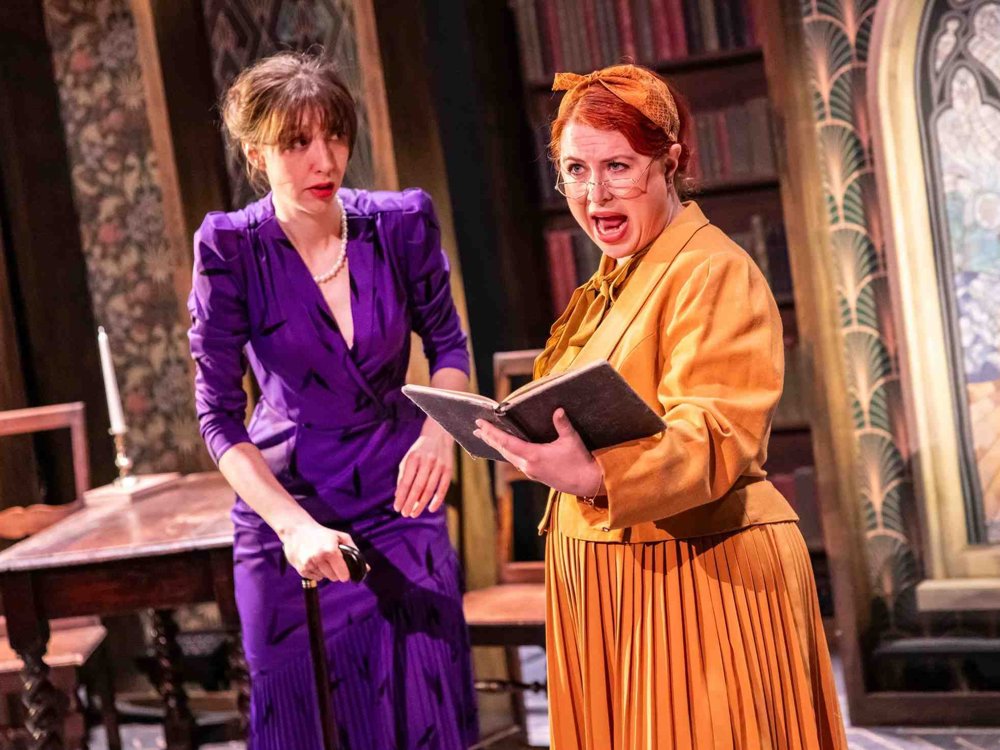 Two performers in colourful costumes; one in a purple dress and pearls holding a cane, and the other in a mustard yellow outfit reading passionately from a book.