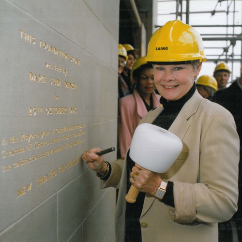 A photograph of actress Dame Judi Dench wearing a hard hat and holding a large mallet, laying the ornamental foundation stone of the building in 1994.