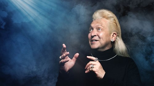 A man with long blond hair and multiple rings gestures expressively while looking upwards. He wears a black turtleneck and silver jewellery, with dramatic lighting and smoky blue background creating a mystical atmosphere