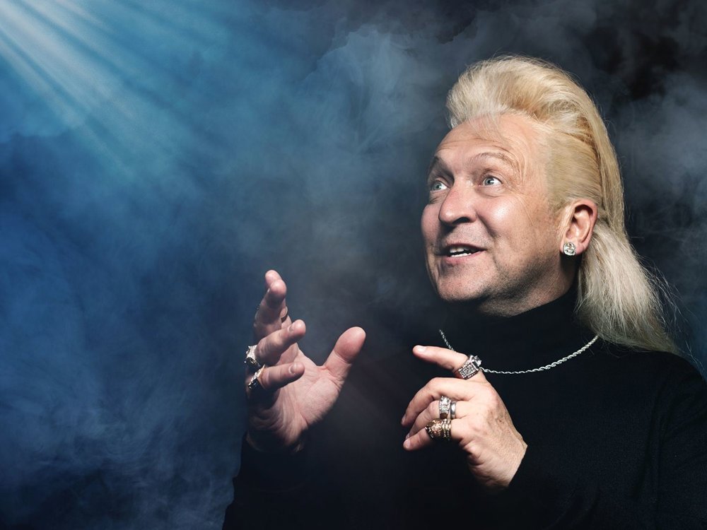 A man with long blond hair and multiple rings gestures expressively while looking upwards. He wears a black turtleneck and silver jewellery, with dramatic lighting and smoky blue background creating a mystical atmosphere
