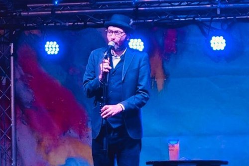 A comedian wearing a dark suit, bowler hat, and glasses stands on an indoor stage holding a microphone. Blue stage lights and a colourful abstract backdrop create a vibrant atmosphere. A drink rests on a small table beside him.