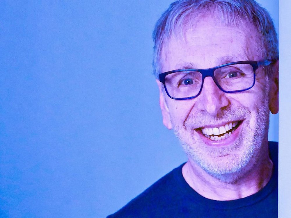 Smiling man with glasses and a grey beard, wearing a dark shirt, peeking playfully from behind a white wall against a blue background. A 'Comedy' badge appears in the top left corner.