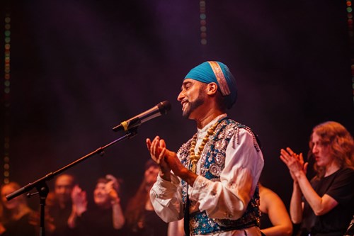 A man in traditional Bhangra costume stands in front of a microphone looking out to the left of the picture from a stage with people behind him. Everyone is clapping.