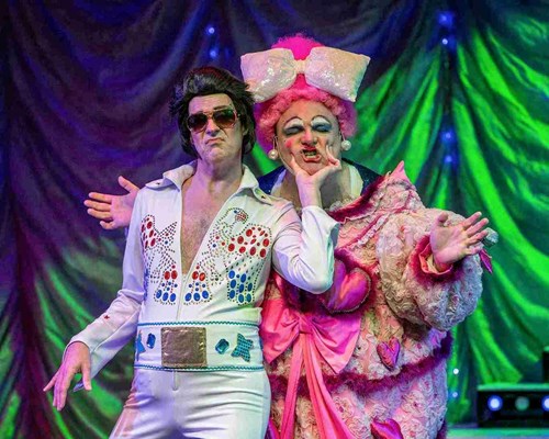 A comedic stage scene featuring a performer dressed as Elvis Presley in a dazzling white jumpsuit adorned with colourful rhinestones and a large metallic belt, complete with oversized sunglasses and an exaggerated pout. Beside them, a flamboyant character in an over-the-top pink costume with heart-shaped details, a massive glittery bow, and dramatic makeup strikes a humorous pose, squishing Elvis's cheeks.