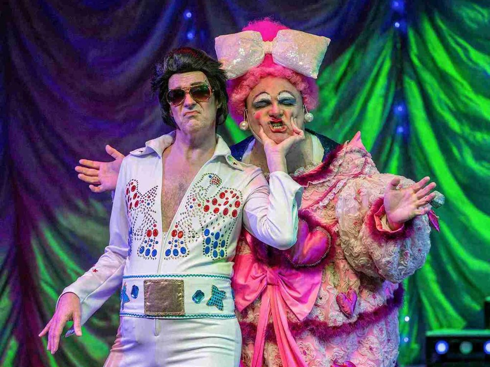 A comedic stage scene featuring a performer dressed as Elvis Presley in a dazzling white jumpsuit adorned with colourful rhinestones and a large metallic belt, complete with oversized sunglasses and an exaggerated pout. Beside them, a flamboyant character in an over-the-top pink costume with heart-shaped details, a massive glittery bow, and dramatic makeup strikes a humorous pose, squishing Elvis's cheeks. 