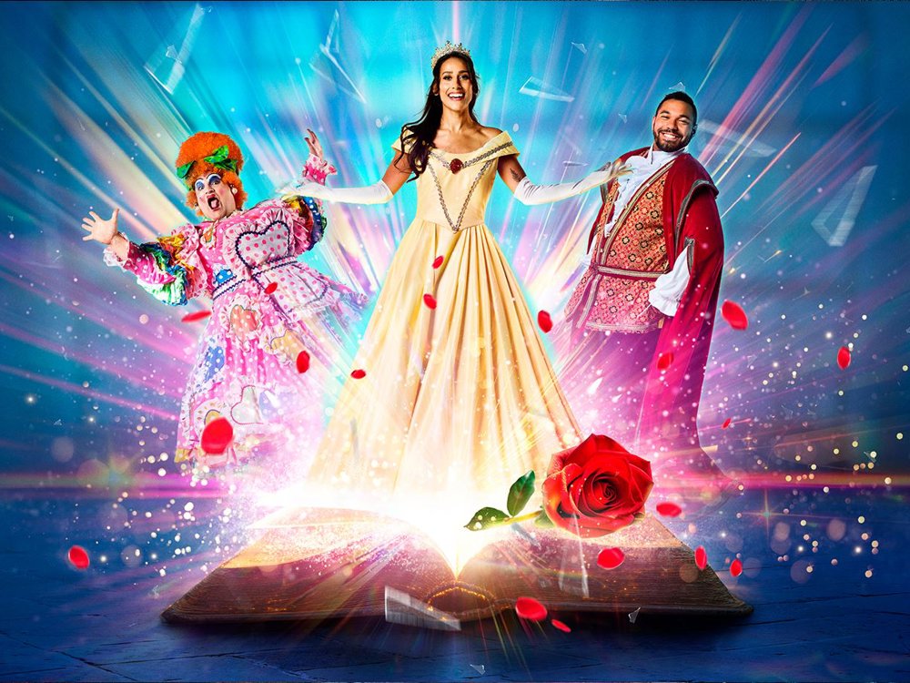 Colourful promotional image for a theatrical production of Beauty and the Beast. A glowing storybook opens at the bottom, emitting sparkling light and rose petals. In the centre stands a smiling woman dressed as Belle in a golden gown and tiara. To her left is a flamboyantly dressed panto dame in a brightly coloured costume and orange wig. To her right stands a man in an ornate red and gold princely outfit. 