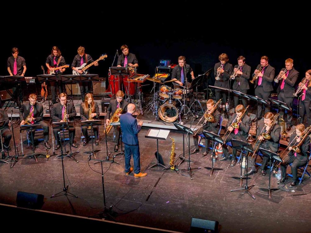 A large jazz band performing on stage, featuring musicians playing brass, woodwind, guitar, bass, drums, and percussion instruments, all dressed in black with pink ties, led by a conductor.