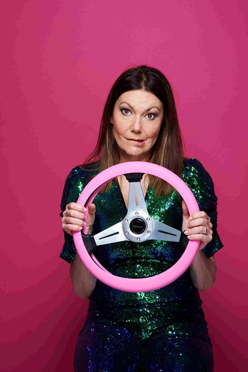 Comedian Fiona Allen has shoulder length brown hair and is wearing a blue sequin dress, she poses holding a pink steering wheel.