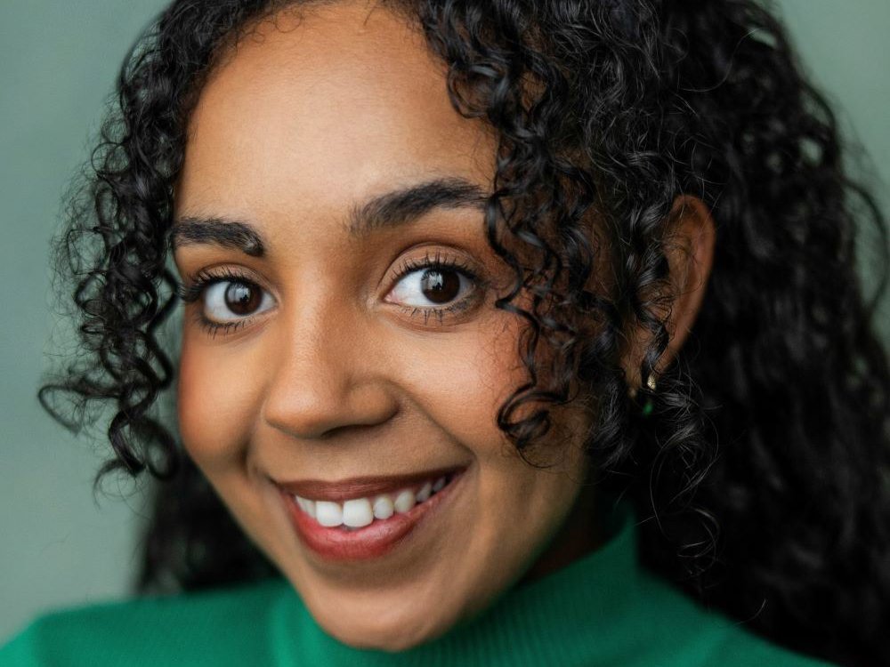 Headshot of Alyce Liburd. She has dark curly hair, wears a green turtleneck, and smiles warmly at the camera.