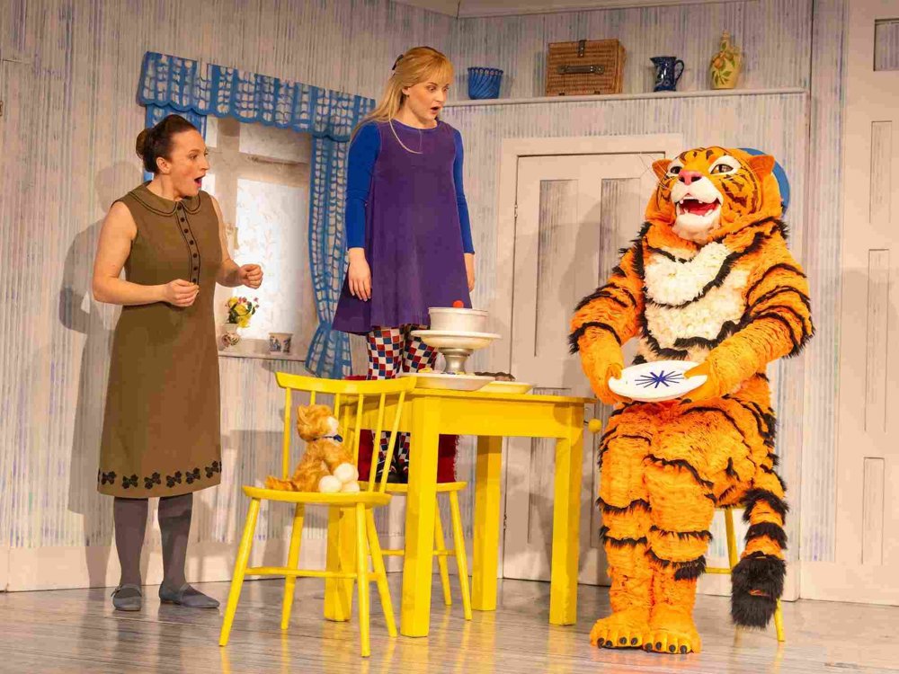 Two women stand in shock beside a yellow table as a large tiger character sits holding a plate, on a colourful vintage-style kitchen stage set.