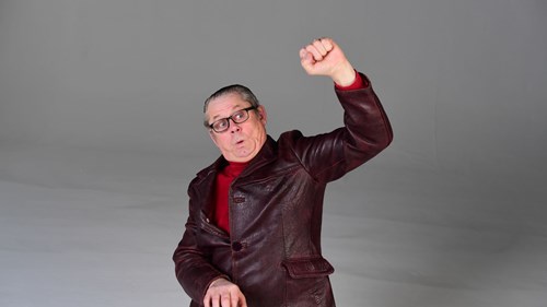 A middle-aged man with slicked-back grey hair and black-rimmed glasses is wearing a burgundy leather jacket over a red turtleneck. He has a surprised or enthusiastic expression, with his mouth slightly open and eyebrows raised. His right arm is raised with a clenched fist, and his left hand appears to be mimicking playing a keyboard. The background is a neutral grey studio setting.