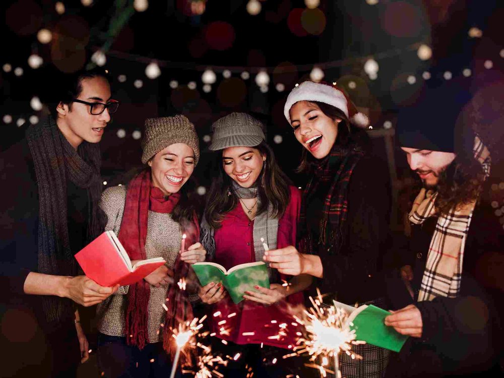A group of five friends stands outdoors, cheerfully singing Christmas carols together. They are dressed warmly in winter attire, with scarves, hats, and coats. The scene is festive and joyful, illuminated by glowing sparklers they hold in their hands. Each person also holds a songbook.