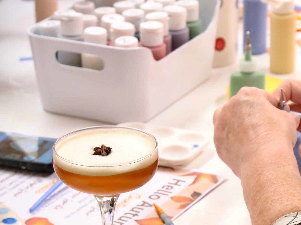 A frothy amber cocktail garnished with star anise sits in a coupe glass on a craft workshop table, surrounded by colourful paint bottles, brushes and art supplies as participants enjoy a painting session.