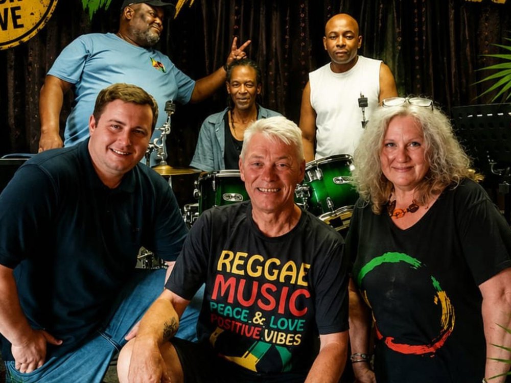 Six people pose together in a music venue with a drum kit and keyboards in the background. Three people stand at the back, while three sit at the front, smiling at the camera. Several wear reggae-themed clothing, and tropical leaf graphics frame the image, creating a vibrant, community-focused atmosphere.