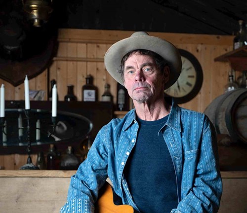 Comedian Rich Hall wearing a beige cowboy hat and navy blue t-shirt with a light blue open shirt. 